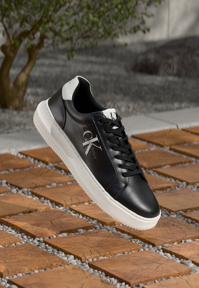 Zwarte leren sneaker met witte rubberen zool, voorzien van vetersluiting en merkreferentie op de zijkant. Achtergrond bevat steen- en houttexturen.
