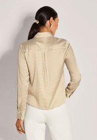 Frau mit dunklem Haar im Pferdeschwanz, die goldene Ohrringe, ein langärmliges Hemd in beige-weißer Hahnentrittmuster und weiße Hose trägt und von hinten zu sehen ist.