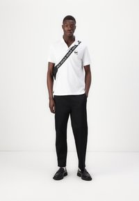 Biely polo tričko s logom, čierna crossbody taška so značkou, čierne nohavice a čierne topánky s hrubou podrážkou, stojí na svetlom pozadí.