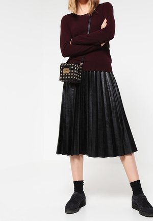 Kvinde iført en burgunderrød langærmet top, sort plisseret midi-nederdel, sorte plateau-sko og en ternet tværs skuldertaske, stående med armene over kors.