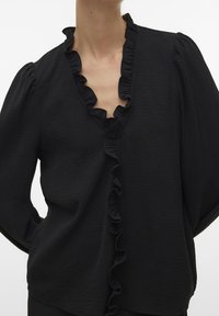 Haut noir en tissu texturé avec un décolleté en V profond. Il présente des manches bouffantes et des volants le long de l'ouverture avant.