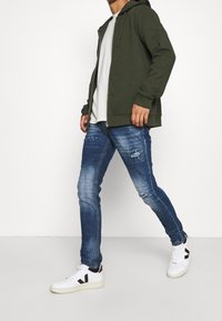 Zelenohnědá mikina na zip s kapucí, kombinovaná s potrhanými modrými džíny a bílými teniskami s černými akcenty.