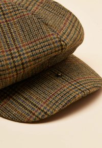 Kapa iz tvida z rjavozelenomodrim karo vzorcem, okrogle oblike, z mehko teksturo in črnim gumbom na sponki.