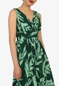 Grünes Blumenkleid mit V-Ausschnitt, gefaltetem Rock und kontrastierendem, hellgrünem Blattmuster. Glatte Textur, taillierter Oberteil und fließendes Design.