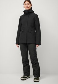 Zwart geïsoleerd jack en broek set, met een hoge kraag, meerdere zakken en een gladde, matte afwerking. Waterdichte schoenen ronden de outfit af.