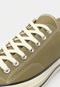 Basket verte en toile avec lacets blancs, œillets en argent et couture blanche contrastante. Dispose d'une semelle en caoutchouc noire et d'une texture lisse.