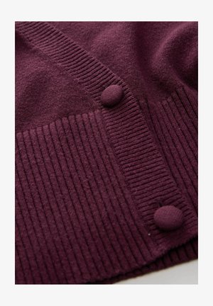 Pletený kardigán v hlbokej fialovej farbe, ktorý má rebrovanú štruktúru a gombíky potiahnuté tkaninou. Hladká horná časť prechádza do textúrovaného spodného dielu.