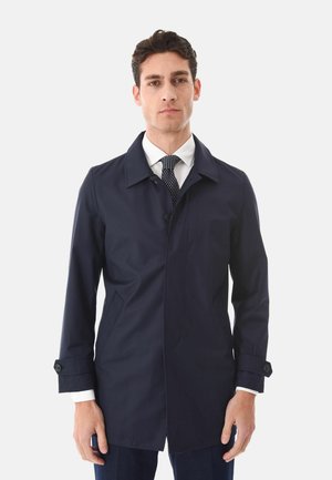 Uomo che indossa un cappotto blu navy con bottoni sopra una camicia bianca e una cravatta nera a pois, in piedi davanti a uno sfondo chiaro e semplice.