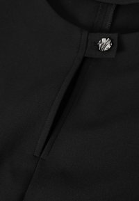 Vêtement en tissu noir à texture lisse, avec une fermeture à boutons et une fente au niveau de l'encolure pour rehausser le design.