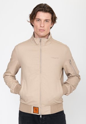 Mann trägt beigen Bomberjacke mit Reißverschlusstasche am Ärmel, steht mit den Händen in den Taschen vor einfachem Hintergrund.