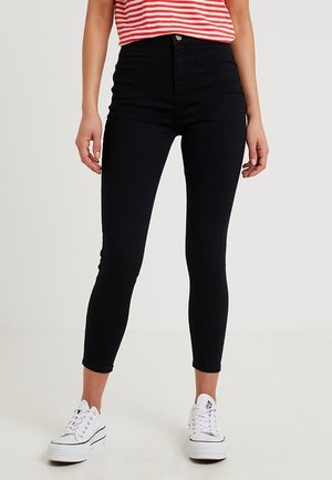 Person, die schwarze High-Waist Skinny-Hosen, ein rot-weiß gestreiftes Shirt und weiße flache Sneaker trägt, steht vor einem einfarbigen Hintergrund.