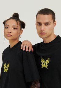 Des t-shirts en coton noir avec un graphique de papillon au motif de flamme jaune sur la poitrine, portés par deux personnes sur un fond neutre.