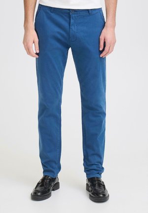 Man wearing straight-fit blue cotton trousers and black leather loafers, standing against a plain background.