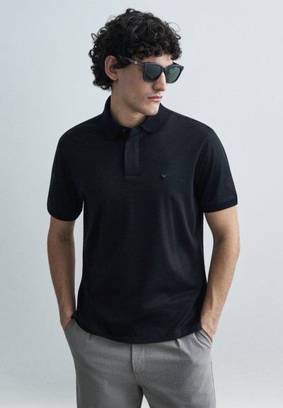Zwart poloshirt met korte mouwen, getextureerde kraag, subtiel logo op de borst, gecombineerd met grijze broek, model draagt donkere zonnebril.