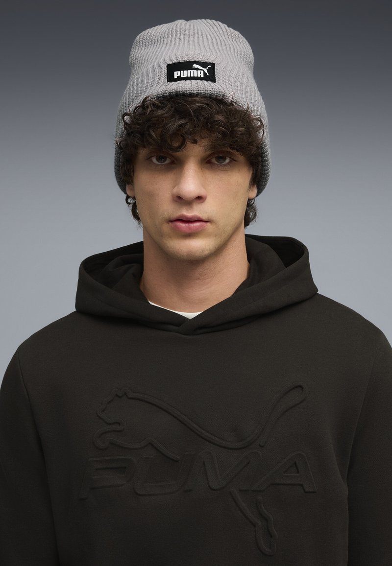 Bonnet en tricot côtelé gris avec un patch logo Puma noir, porté avec un sweat à capuche noir arborant un design Puma en relief.