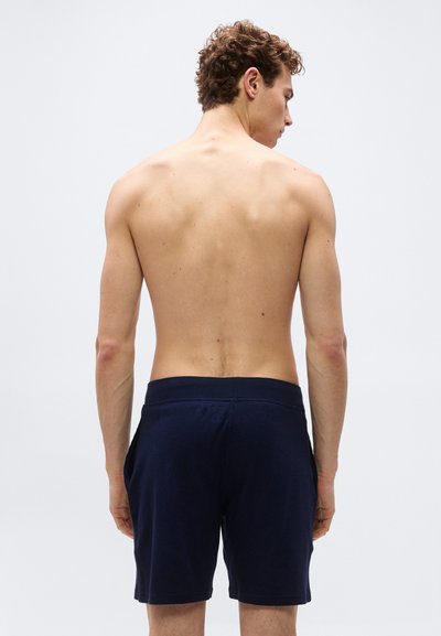 Νέος άνδρας με σγουρά μαλλιά, χωρίς μπλούζα, φορώντας σκούρα μπλε ναυτικομπλε σορτς, όρθιος με την πλάτη προς τα εμπρός σε απλό ανοιχτό γκρι φόντο.