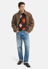Bruine, gestructureerde jas over een multicolor argyle trui, gecombineerd met lichtblauwe jeans en zwarte schoenen. Het ontwerp heeft een hoge kraag.