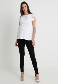 Blusa branca com mangas em babados e corpete texturizado, combinada com jeans skinny pretos e sapatos de salto abertos brancos. Fundo neutro.
