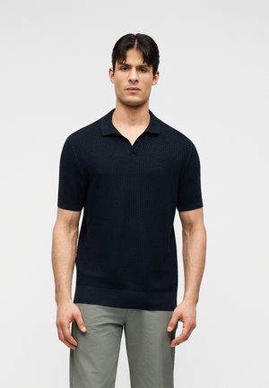 Hombre vistiendo una camisa tipo polo ajustada de manga corta texturizada en azul marino y pantalones gris claro, de pie frente a un fondo liso de color gris claro.