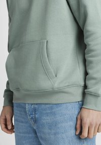 Haut de sweat à capuche en coton vert avec une poche sur le devant, des poignets côtelés et une couleur unie. Associé à un jean en denim bleu clair.