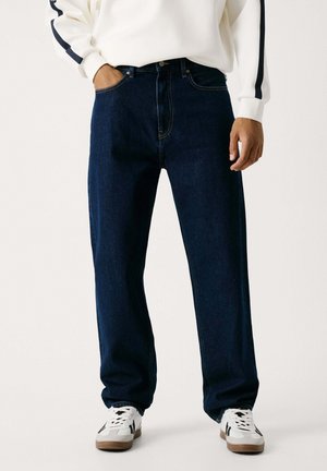 Jeans in denim blu scuro con vestibilità dritta, dotati di cinque tasche, cuciture a contrasto e un design classico per le passanti della cintura. Abbinati a sneakers bianche.