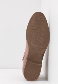 Semelle d'une chaussure marron clair montrant un motif de bande de roulement texturé, taille 37 indiquée, appuyée contre une surface d'angle blanche.