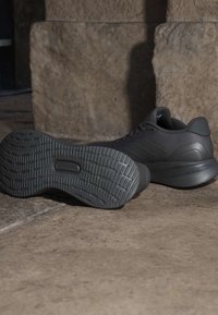 Pair of black athletic shoes, one showing detailed textured sole, placed on stone floor against a stone wall background.