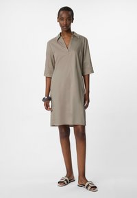 Khakifarbenes Kleid mit Kragen, Dreiviertelärmeln und V-Ausschnitt. Das Modell trägt schwarze und beige Rutsch-Sandalen. Glattes Material, lockere Passform.