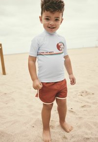 Modrý rash guard s krátkým rukávem a červenozeleným logem, doplněný terakotovými plavkami s bílým lemem. Pozadí tvoří písčitá pláž.