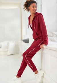 Bordeauxfarbene Kapuzenjacke mit Reißverschluss und Kordeln, kombiniert mit passenden Jogginghosen mit weißen Seitenstreifen. Getragen mit weißen Sneakern. Glatte Textur.