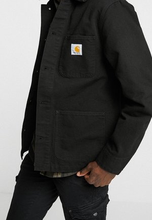 Person trägt eine schwarze Carhartt-Jacke mit Fronttaschen und schwarze Cargohosen, Hand ruht nahe der Tasche, vor einem einfarbigen hellen Hintergrund.