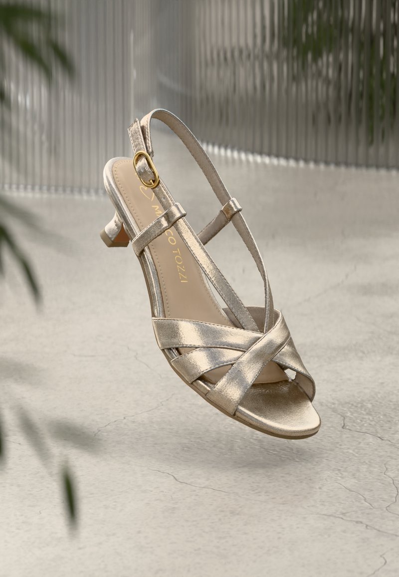 Zlatne metalik sandale s otvorenim prstima, s ukriženim trakama, kopčanjem na peti i niskim kitten potpeticama na teksturiranoj betonskoj površini.