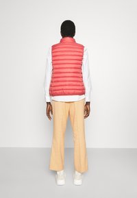 Red puffer vest with horizontal stitching, high collar, worn over a white shirt and light beige wide-leg pants. Smooth texture, casual fit.