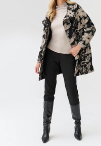 Abrigo floral negro y beige sobre un suéter de cuello alto claro, combinado con pantalones negros y botas de tacón alto negras hasta la rodilla.