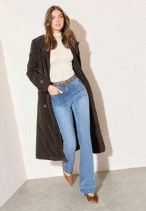 Frau trägt einen langen dunklen Mantel, beigen Rollkragenpullover, blaue Jeans mit Vordertaschen, braunen Gürtel und braune spitze Absätze, steht an einer weißen Wand.