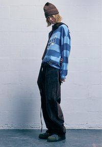 Modrá a námornícky pruhovaná košeľa s dlhým rukávom, tmavé voľné nohavice, zelené topánky s modrými akcentami a hnedý pletený čiapka. Stojí pri stene.