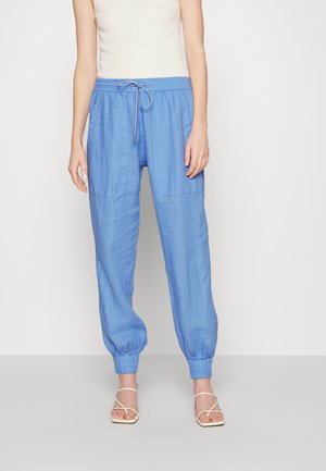 Kobieta ubrana w jasnoniebieskie lniane spodnie typu jogger ze sznurkiem i elastycznymi mankietami oraz białe sandały na paski, stojąca na jednolitym tle.