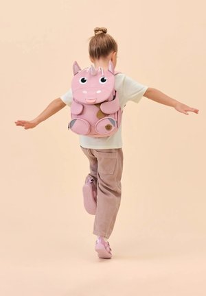 Child with hair in a bun, wearing a white shirt and pink pants, balancing on one foot while carrying a pink unicorn backpack.