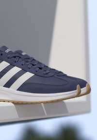 Zapatillas Adidas azul marino con rayas blancas y suelas de goma descansando sobre una superficie blanca con un fondo neutro.