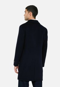 Man in een donkerblauwe lange jas, staande met zijn rug naar de camera tegen een effen lichte achtergrond.