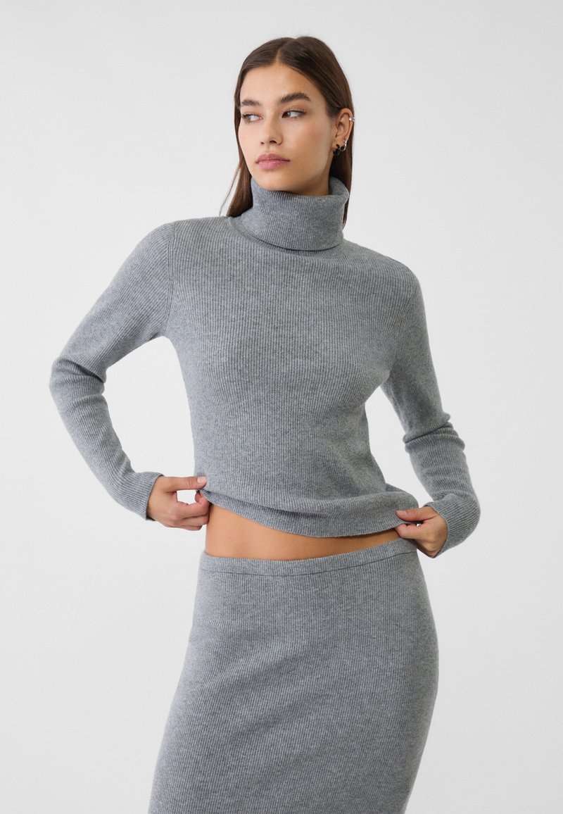 Frau, die einen grauen, gerippten Rollkragenpullover anpasst, trägt einen passenden gerippten Rock und steht vor einem schlichten hellen Hintergrund.