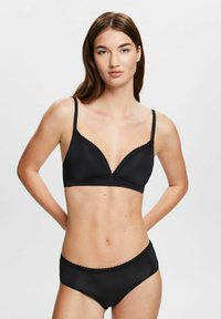 Woman wearing a black bra and matching black panties with lace trim, standing against a plain light gray background.