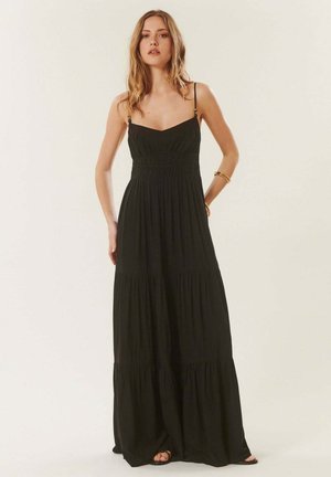 Femme portant une robe longue noire sans manches avec des bretelles fines et une jupe à volants, debout devant un fond clair uni.