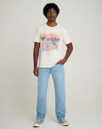 Ανοιχτό καφέ γραφικό t-shirt με ρετρό σχέδιο που περιλαμβάνει το κείμενο "Wrangler", συνδυασμένο με ανοιχτό μπλε tapered παντελόνι και γκρι αθλητικά παπούτσια.