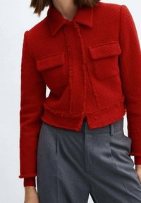 Red cropped jacket in textured fabric, featuring a collar, front button closure, and two front pockets with frayed edges. Paired with gray trousers.