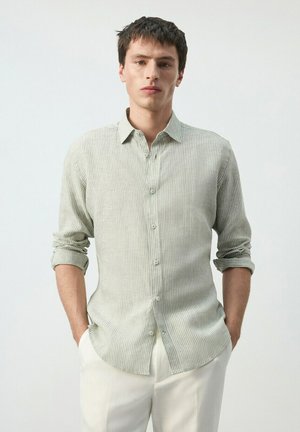 Jeune homme les mains dans les poches, portant une chemise à rayures grise claire boutonnée et un pantalon blanc, sur un fond uni.