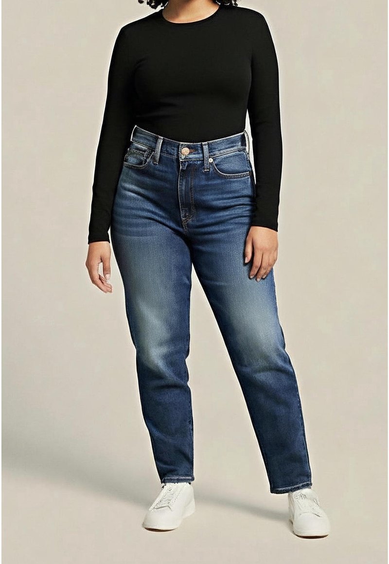 Femme portant un haut noir à manches longues, un jean bleu taille haute et des baskets blanches, debout devant un fond uni.