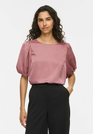 Femme aux cheveux bouclés et foncés portant un chemisier mauve à manches courtes bouffantes et un pantalon noir taille haute, debout avec une main dans la poche sur fond blanc.