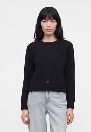 Femme aux longs cheveux noirs portant un cardigan noir boutonné et un jean bleu clair, debout devant un fond blanc uni.