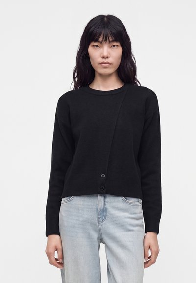 Femme aux longs cheveux noirs portant un cardigan noir boutonné et un jean bleu clair, debout devant un fond blanc uni.